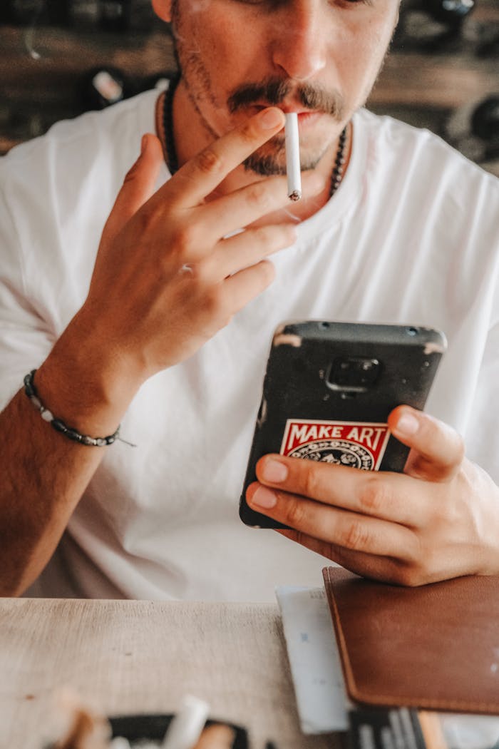 A man in a casual setting smoking a cigarette and using his smartphone, showing a relaxed lifestyle.