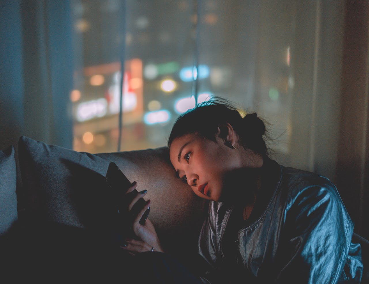 A woman looking at her mobile phone at night with city lights blurred in the background.